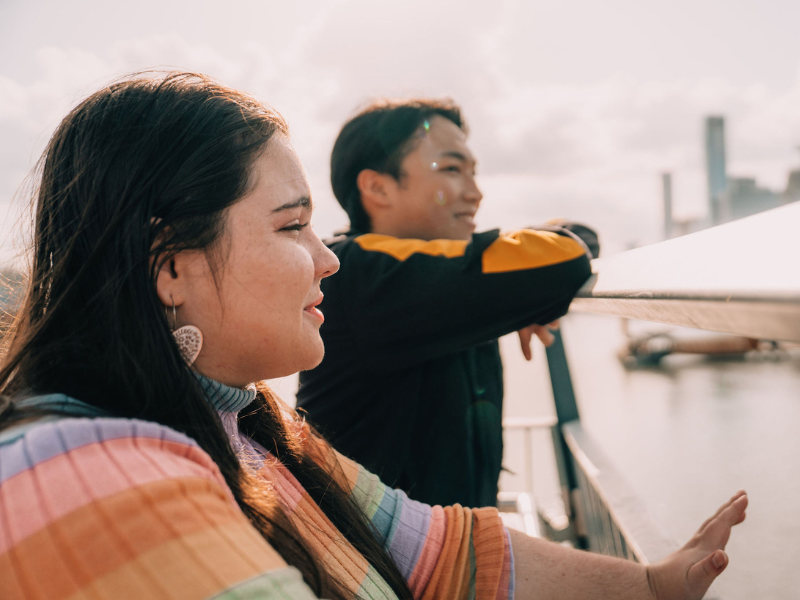 Two people stand on a bridge overlooking water, with city buildings visible in the background on a bright day.