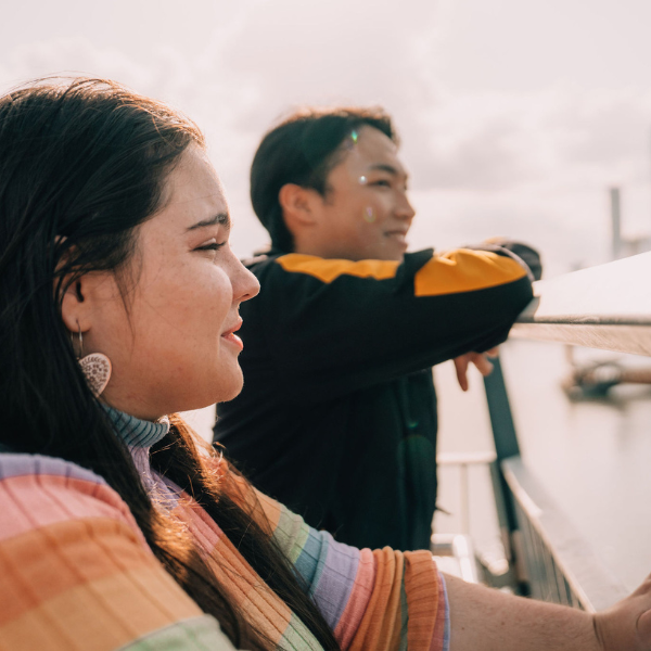 Two people stand on a bridge overlooking water, with city buildings visible in the background on a bright day.
